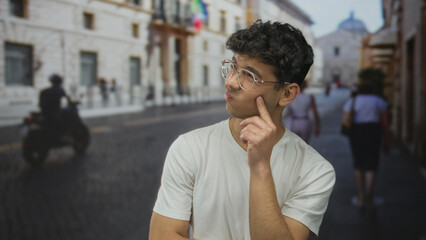 Fototapeta premium Young hispanic man wearing glasses hand on chin thinking on sunlit cobblestone street in historic city center; curiosity.