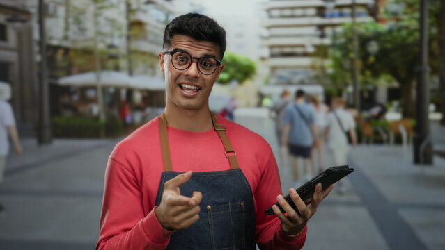 Smiling man with glasses in apron holding tablet outdoors at city terrace cafe with people and buildings in background