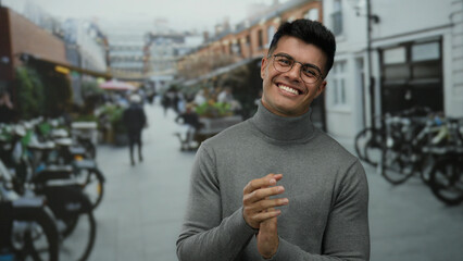 Naklejka premium Young man clapping hands in a lively outdoor city street wearing glasses and a sweater with people and bicycles in the background showcasing a vibrant urban environment.