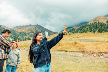 young Asian woman with long black hair takes a selfie in the mountains. Two men and a boy stand nearby, enjoying the scenic view.