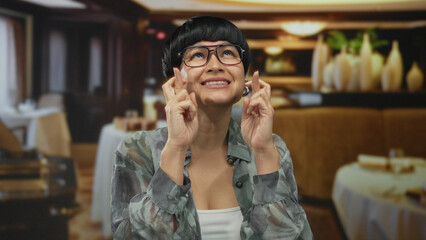 Woman smiling with crossed fingers in a cozy restaurant, wearing glasses and a casual shirt under warm lighting, creating a hopeful and relaxed indoor dining atmosphere.