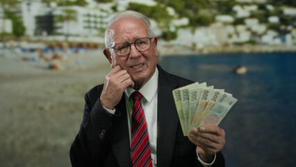 Senior man holding czech koruna banknotes at a seaside beach, wearing a business suit, reflecting on opportunities and prosperity by the water.