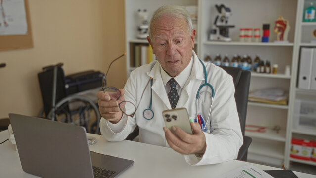 Senior male doctor in clinic examines smartphone near laptop in medical office.