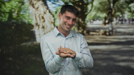 Young hispanic man in lightblue shirt rubs fingers for money gesture in sunlit green forest; anticipation.