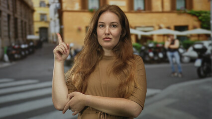 Woman pointing finger upward on street beneath urban building facade with visible freckles and red hair; curiosity.
