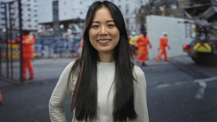 Fototapeta premium Young chinese woman smiling at a construction site outdoors with workers in orange safety suits, capturing a blend of human interaction and urban development.