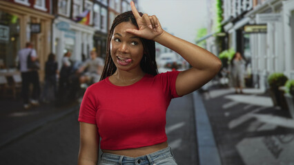 Woman holds l sign on forehead with hand on a busy street wearing red shirt and jeans; playful mischief.