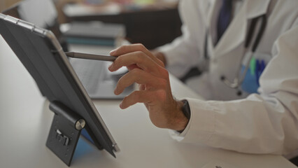 Young hispanic doctor man in white coat taps tablet screen with stylus at clinic desk beside laptop...