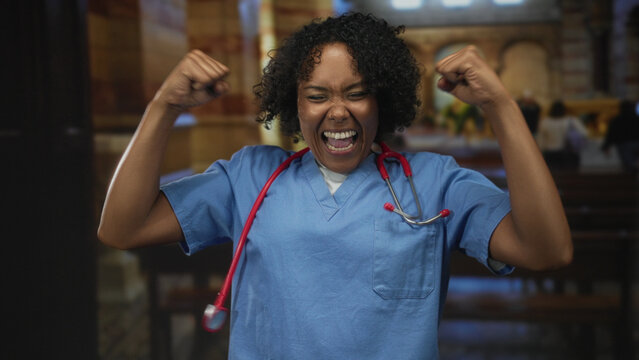 Young african american doctor with stethoscope clenches fists and shouts inside church building aisle; elation.