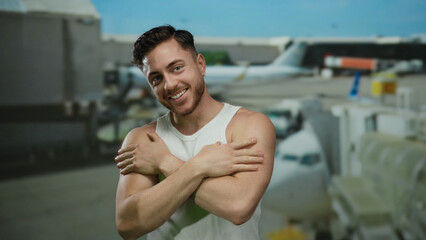 Fototapeta premium Young man with beard smiling joyfully at an airport terminal embracing himself with airplanes in the background.
