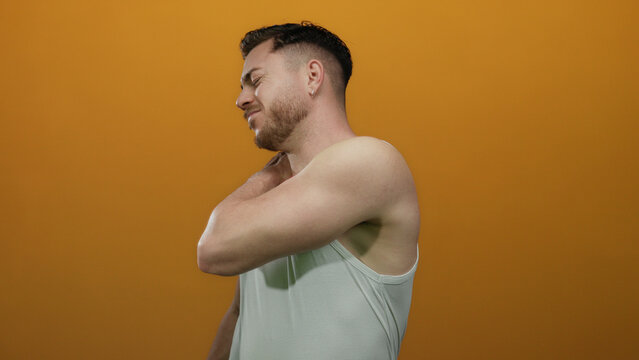 Young man with beard in white tank top experiencing shoulder pain against isolated orange background, conveying discomfort and health theme. - Powered by Adobe