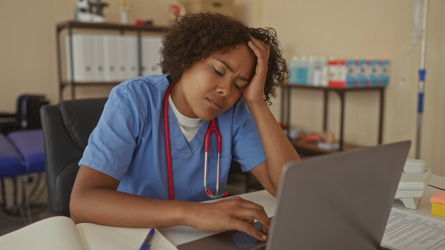 Young african american woman doctor typing on laptop at clinic desk furrows brow; stress fatigue overload.