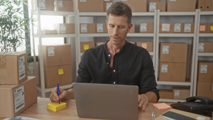 Young man waves hand at laptop while packing parcels on desk with cardboard boxes on shelves in building; focus.