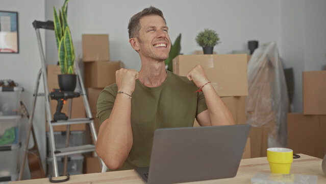 Man raising fists and smiling over laptop in building with moving boxes and ladder seated at wooden table; celebration.