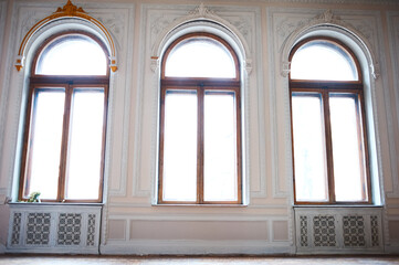 Bright light pours through three tall arched windows set in a historic, intricately decorated room. Soft pastel walls, carved details, and lattice panels create a serene, timeless atmosphere.