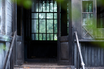 Weathered black wood porch with open doors invites exploration. Soft natural light filters through large panes, revealing a calm, secluded entryway framed by greenery and a rustic atmosphere.