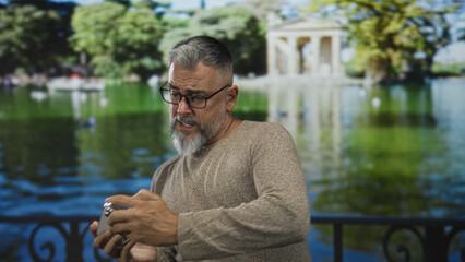 Man holding smartphone with both hands by a lake railing near a building, wearing glasses and a beige sweater; mild frustration.