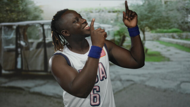 Man in usa basketball jersey number 6 pointing finger upward with both hands and smiling in street near golf cart; joyful confidence.