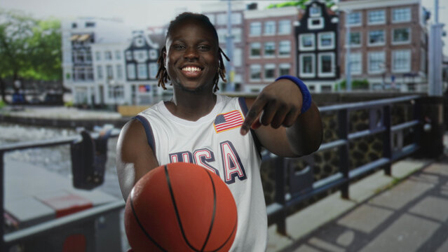 Man points finger to basketball on street beside canal guardrail, smiling with wristband and usa jersey; youth sports joy.