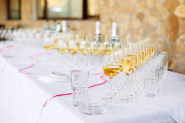 A long white table is lined with numerous champagne and water glasses, pink ribbon accents, and blurred bottles in the background, conveying celebration, hospitality, and refined party atmosphere.