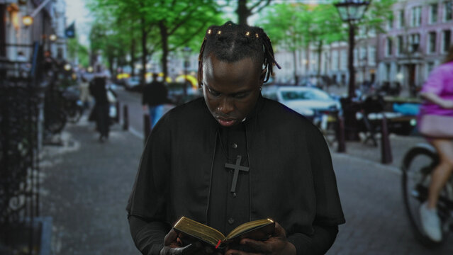 Man reading bible on a busy city street, holding open book and wearing clerical collar and cross necklace; contemplation faith.