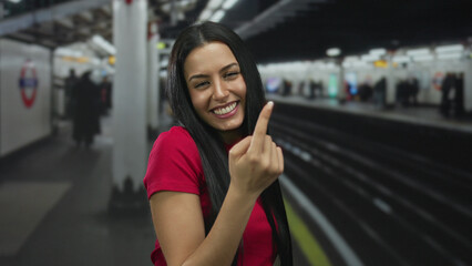 Hispanic woman in a red shirt smiling and gesturing at an indoor train station with blurred railway and commuters in the background.