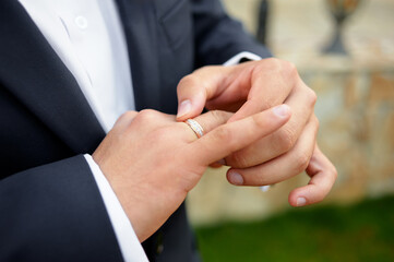 Obraz premium Close-up of hands in a dark suit adjusting a diamond ring on a partner's finger, capturing commitment and romance in a stylish, outdoor wedding moment with a soft background.
