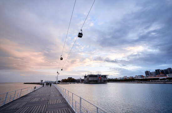 A scenic view of a waterfront with a cable car line, modern buildings, and a wooden walkway extending into the water under a cloudy sky.