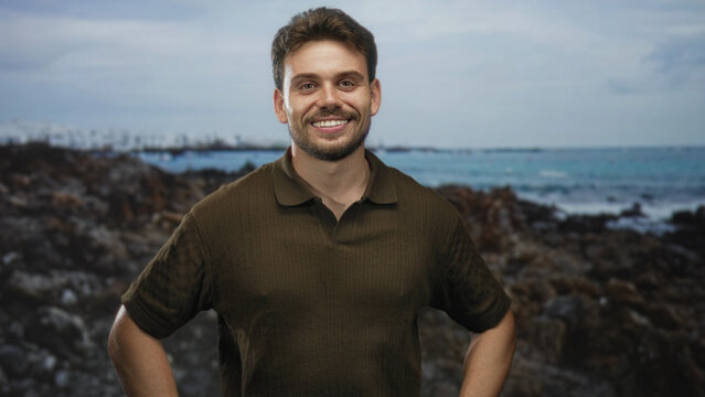 Man with hands on hips smiling toward the sea on rocky coast wearing a brown polo shirt with rolled sleeves; serenity leisure.