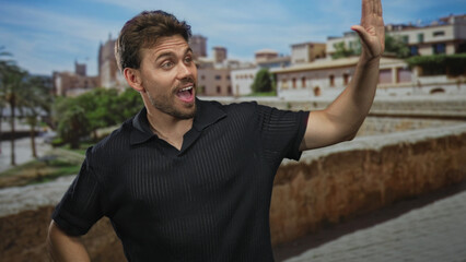 Fototapeta premium Man waving right arm on old town street next to stone wall, smiling and looking aside while leaning slightly; joyful travel moment.