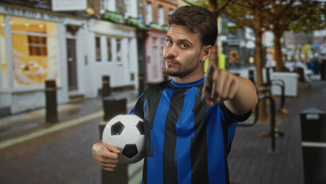 Man holding soccer ball and pointing finger at camera on street in city wearing blue jersey; confidence.