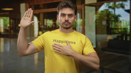 Fototapeta premium Young hispanic man with raised right hand and left hand on chest, wearing yellow t shirt that reads happiness, standing in a hotel building lobby with couches and plants; honor duty.