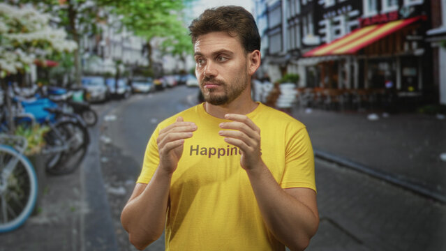 Young man clapping hands in a yellow t shirt on a city street lined with bicycles and outdoor cafe tables, hands held near chest; thoughtful reflection.