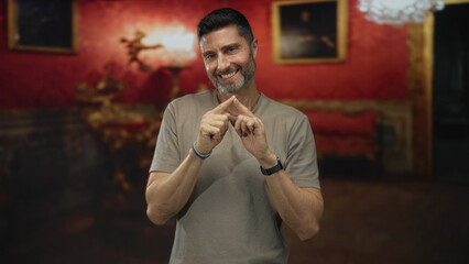 Man smiling touching index fingers together with both hands in ornate museum gallery with red walls and framed paintings; playful warmth.