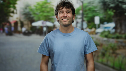 Smiling man in blue shirt stands outdoors on sunny urban street with trees and blurred buildings, exuding friendliness and confidence.