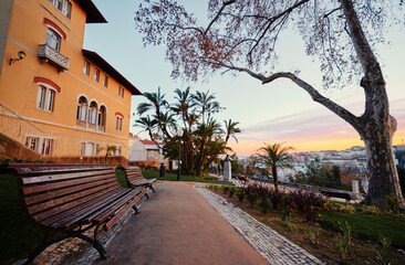 A scenic view of a park with a bench, trees, and a historic building at sunset. The sky is colorful, and the cityscape is visible in the background, creating a tranquil atmosphere.