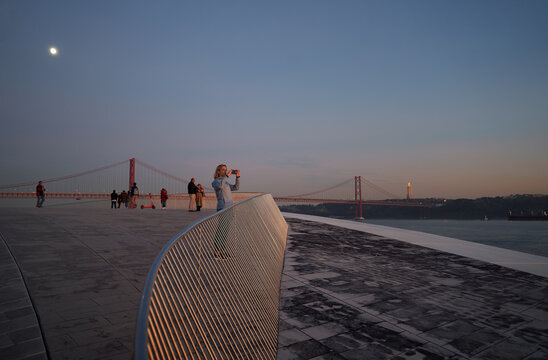 A person taking a photo at sunset on a modern bridge with a cityscape in the background.