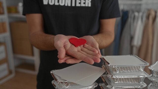 Hispanic man holding red heart symbol while smiling at camera in volunteer room surrounded by stacks of food trays exemplifying kindness and community support
