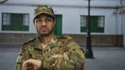 Soldier in camouflage uniform checks watch on wrist while standing on an empty old town street beneath a lamp post; discipline duty.
