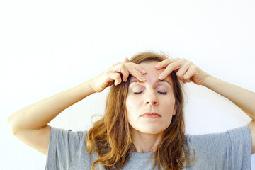 Morning skincare routine: a woman standing against a clean white background gives herself a massage by working her fingers across her forehead. Self-care habit formation and facial yoga practice.