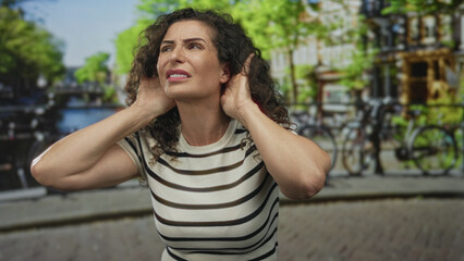 Naklejka premium Woman lifts hand to ear by canal street to listen to ambient noise and navigate the busy waterside path; curiosity.