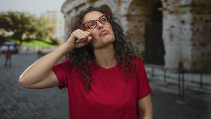 Young hispanic woman outdoors rubs eyes for crying at ancient roman coliseum building; sadness...