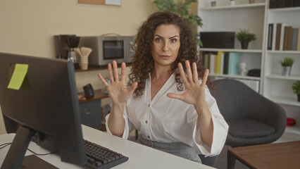 Obraz premium Woman in white shirt raises palms at computer on office desk, showing bare arms; caution and steady focus.