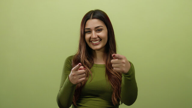 Woman smiling with long hair pointing forward against a vibrant green background, showcasing confidence, positivity, and youthful energy in an isolated studio setting.
