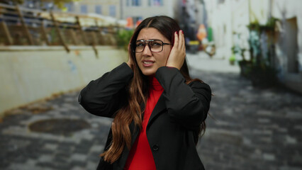 Obraz premium Woman in frustration covering ears on a city street wearing glasses and black jacket outdoors during daytime.