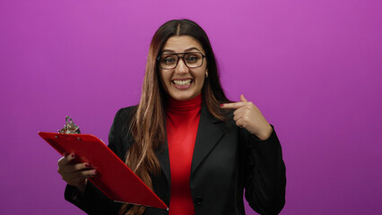 Hispanic woman in glasses pointing to herself confidently holding red clipboard on isolated pink...