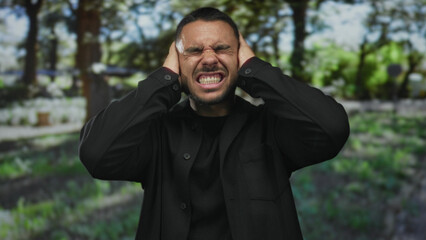 Young hispanic man covers his ears with both hands in sunlit forest clearing under tall green...