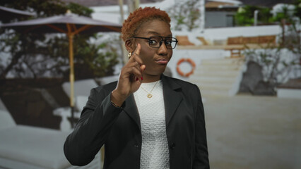 Woman points finger to chest in outdoor building courtyard wearing black blazer and glasses; confidence.