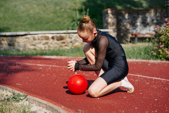 A young girl dressed in an athletic outfit practices rhythmic gymnastics with a red ball on a sunny day outdoors. Young Girl Performing Rhythmic Gymnastics Outdoors Pumping a Red Ball - Powered by Adobe