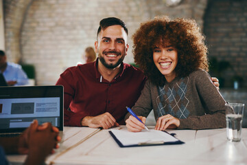 Extremely happy couple smiles directly at the camera while signing a financial document in a consultation meeting